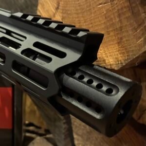 Close-up of the barrel and handguard featuring the Barrel Shroud & Compensator 3″ on a black rifle, set against a wooden background. The image emphasises the vented compensator and the angular, tactical design of the rail system.