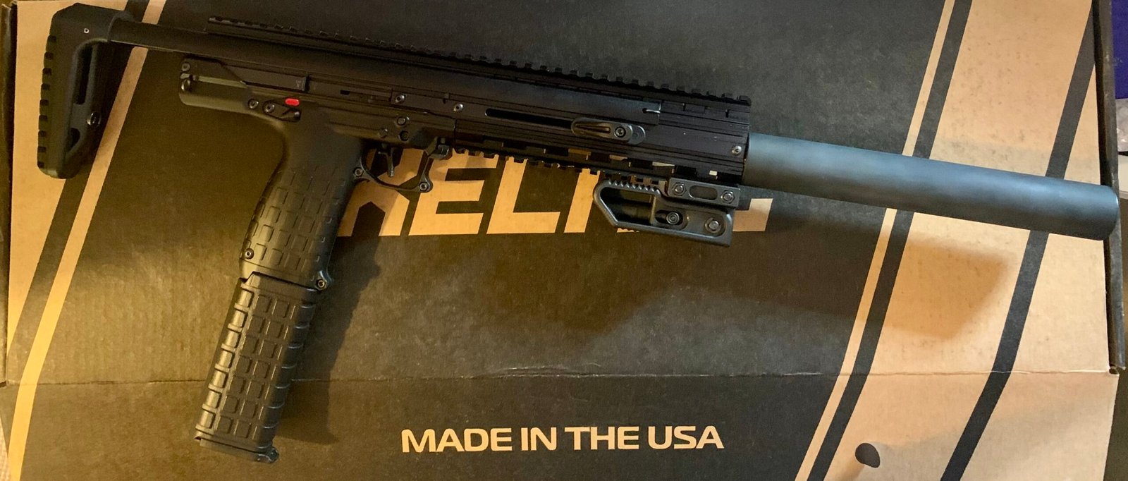 The Keltec CMR30 .22WMR Custom Over-Barrel Moderator, a black semi-automatic firearm with a folding stock and textured grip, is shown on a box labelled MADE IN THE USA in white capital letters.