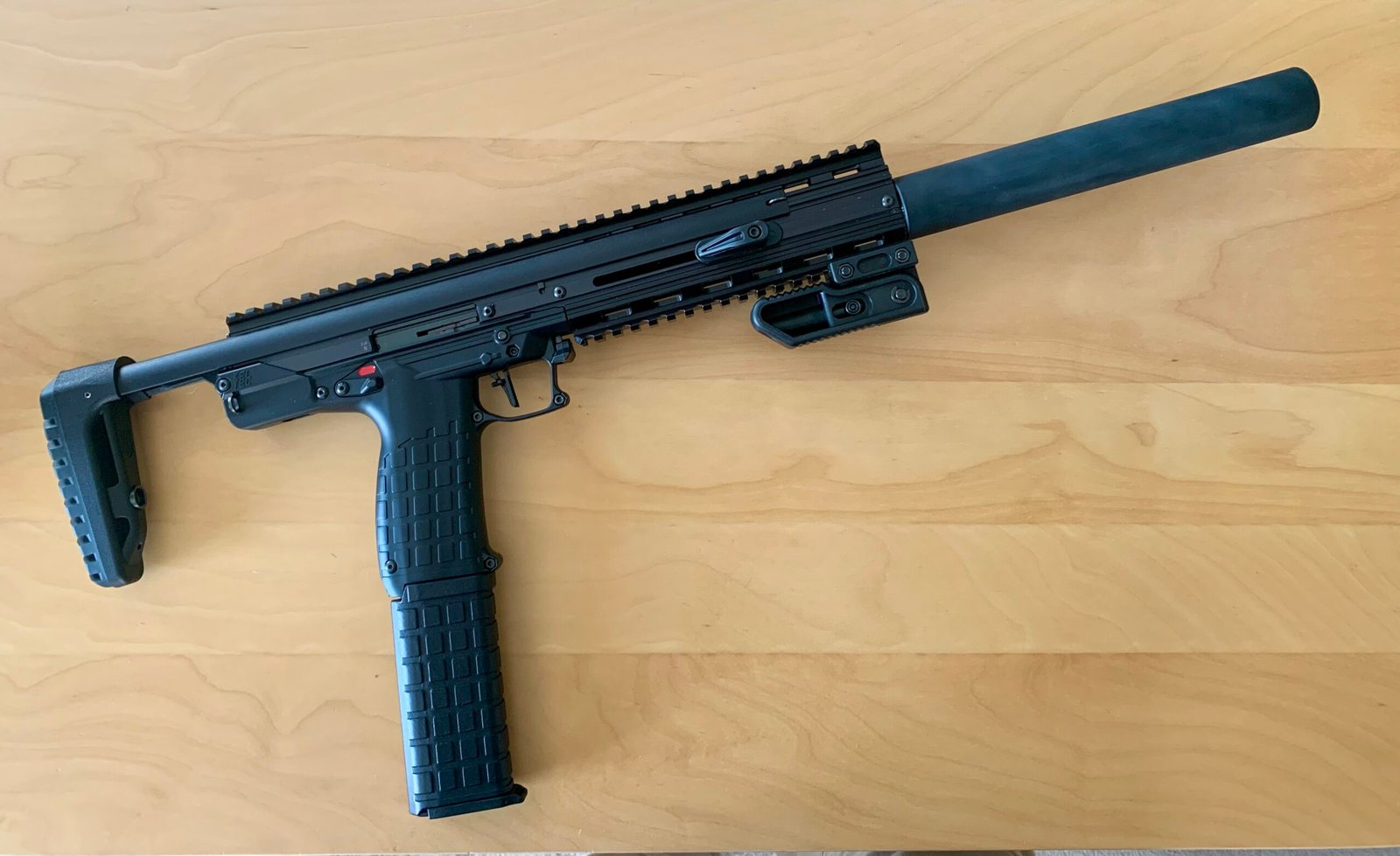 A Keltec CMR30 .22WMR Custom Over-Barrel Moderator with a folding stock, suppressor, rail system, and angled foregrip rests on a light wood surface.