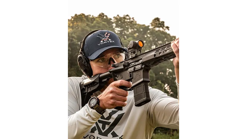 Wearing safety glasses, a cap, and ear protection, a shooter aims a black rifle fitted with the Vortex Triumph 2MOA Red Dot sight outdoors among trees.