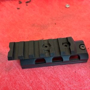 A black metal rail mount with screws, likely for attaching accessories to equipment, lies on a textured red surface.