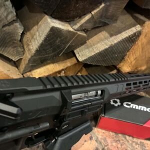 A MAPS-AP22 .22LR AR15 Rifle—fully upgraded and match ready, in black—is positioned in front of a stack of cut firewood, with part of a red and black CMMG-branded box visible beside it.