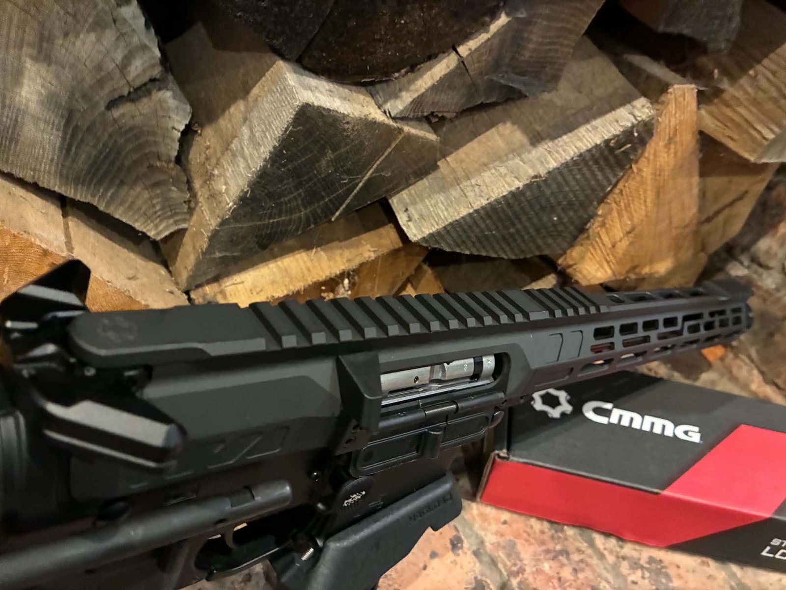 A MAPS-AP22 .22LR AR15 Rifle—fully upgraded and match ready, in black—is positioned in front of a stack of cut firewood, with part of a red and black CMMG-branded box visible beside it.