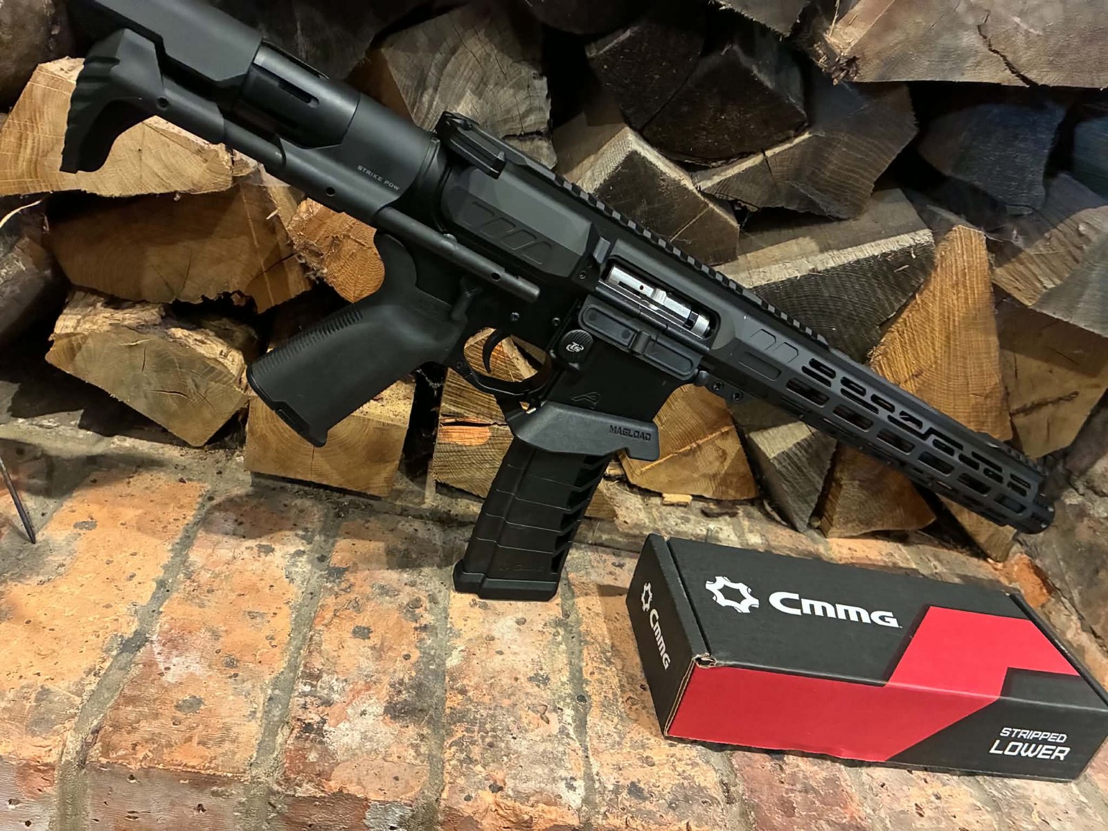 A MAPS-AP22 .22LR AR15 Rifle—fully upgraded and match ready—rests on a brick hearth in front of stacked firewood, with a red and black CMMG box labelled “Stripped Lower” placed nearby.